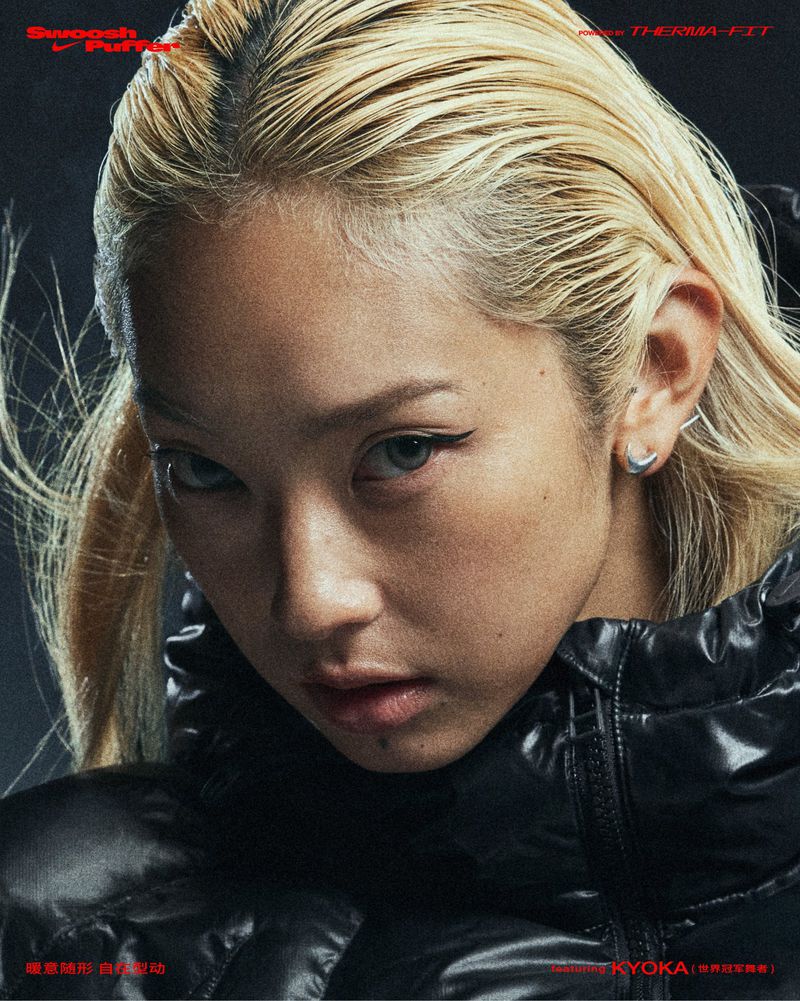 Close-up of a young adult woman with wet blonde hair wearing a black puffer jacket, looking directly at the camera with a serious expression.