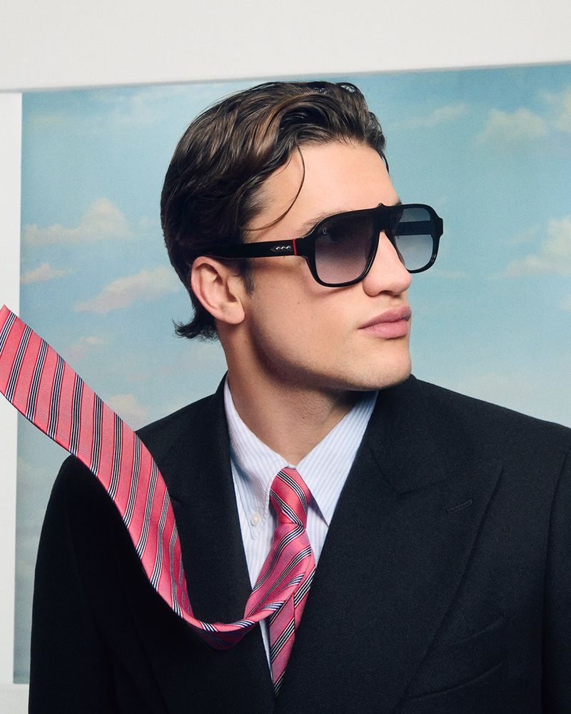 Portrait of a fashionable adult male wearing sunglasses, a white shirt, a black suit, and a red striped tie against a light blue backdrop.