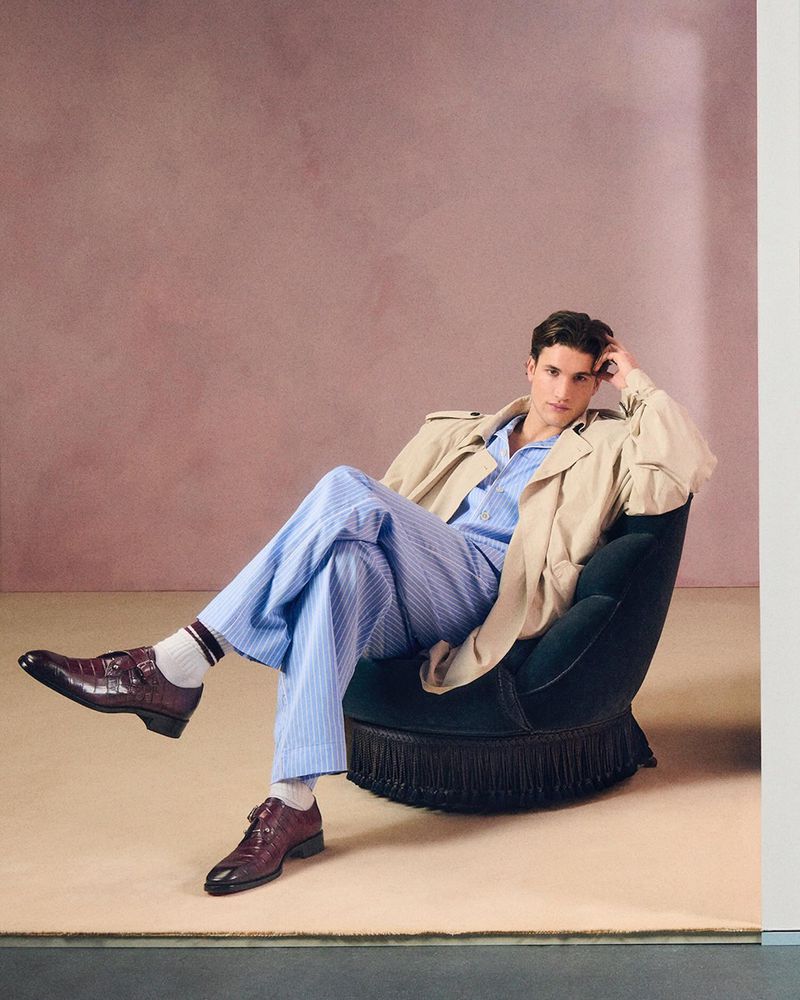 A stylish adult man in a tan trench coat and blue and white striped pajamas is sitting in a green velvet chair in a photo studio.