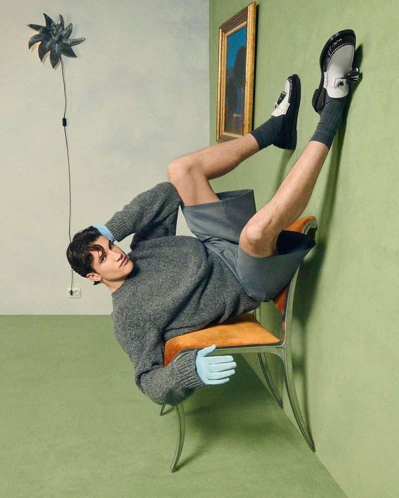 An adult male poses in an indoor fashion setting, wearing a grey sweater, shorts, socks, and loafers. He is in a chair with green walls.