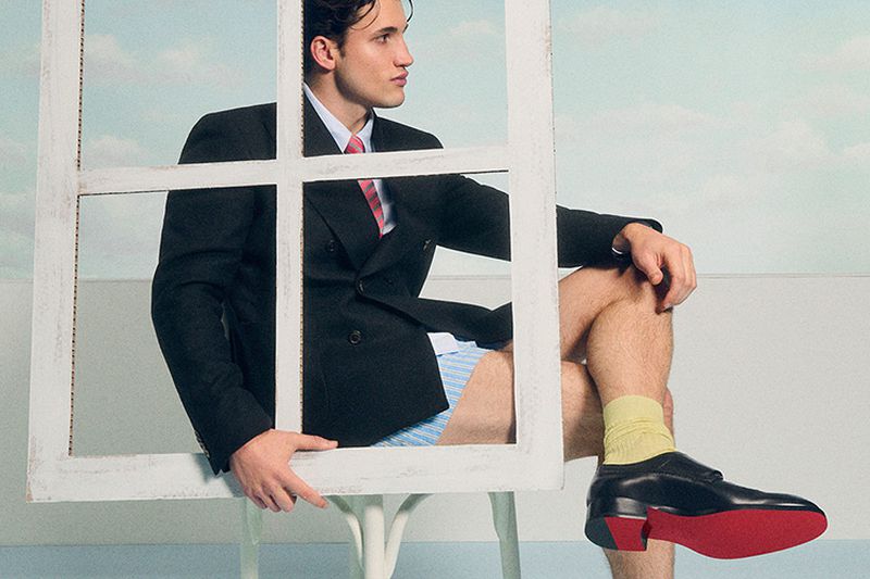 Man in a suit jacket sitting on a chair and holding a white frame in a studio setting, wearing boxer shorts and yellow socks.