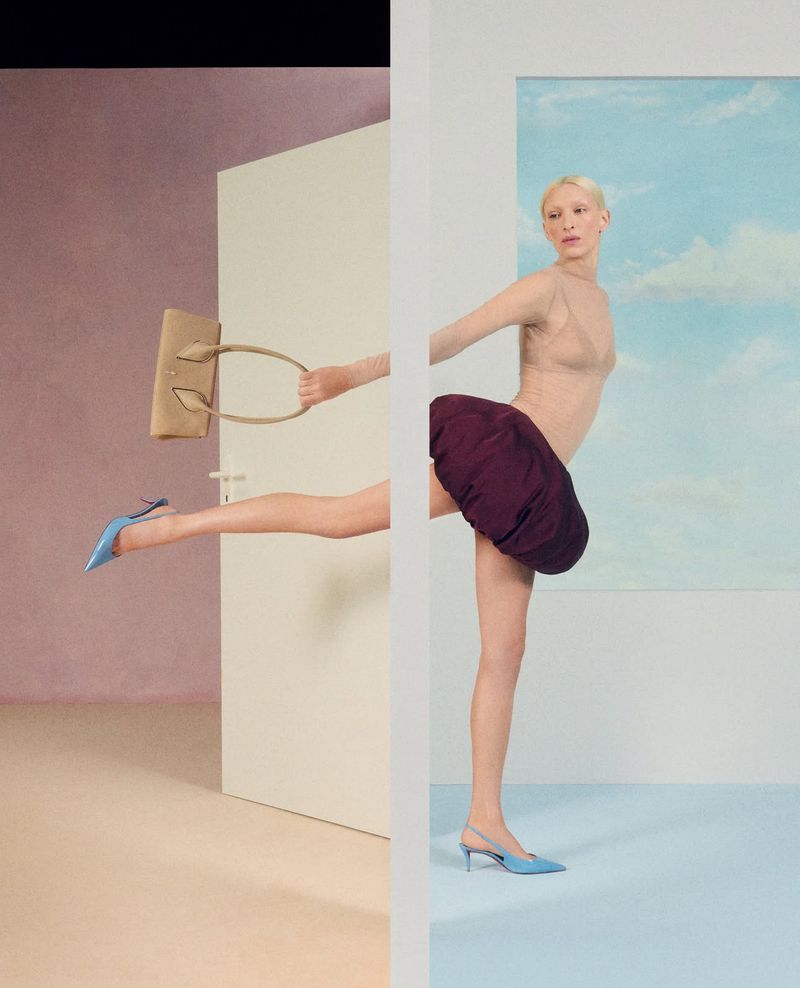 Fashion editorial shot featuring an adult female model holding a tan purse, wearing a purple skirt, and light blue shoes in a studio.