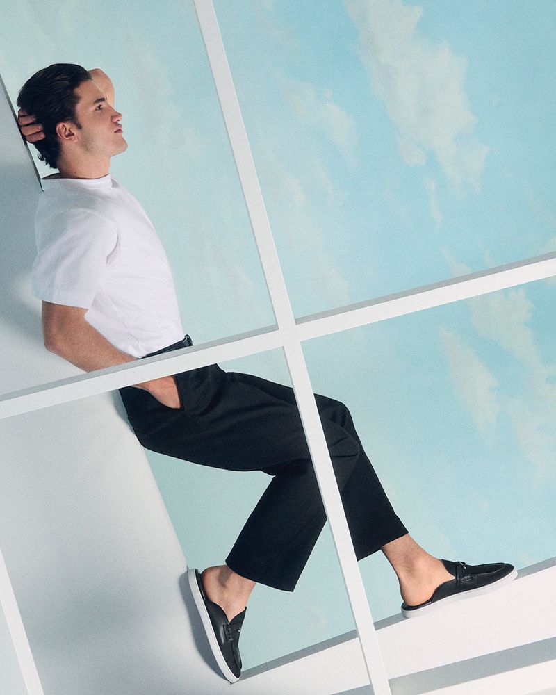Man in white t-shirt and black pants poses against a window with a sky and clouds background, modeling black leather mule shoes.