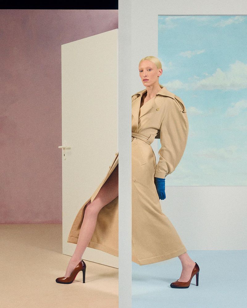 Fashion model in a beige trench coat, wearing blue gloves and heels, poses near a doorway with a blue sky backdrop.