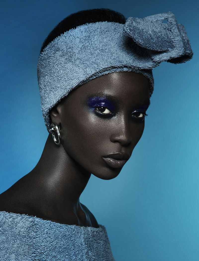 Close-up portrait of an African woman with blue eye makeup and a matching blue towel headband and top, set against a vibrant blue background.