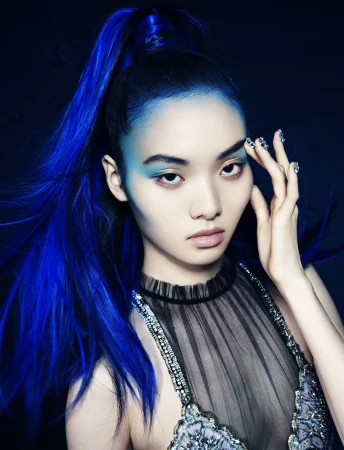 Glamour shot of a model with blue hair and striking makeup, wearing a sheer top with embellishments against a dark background.