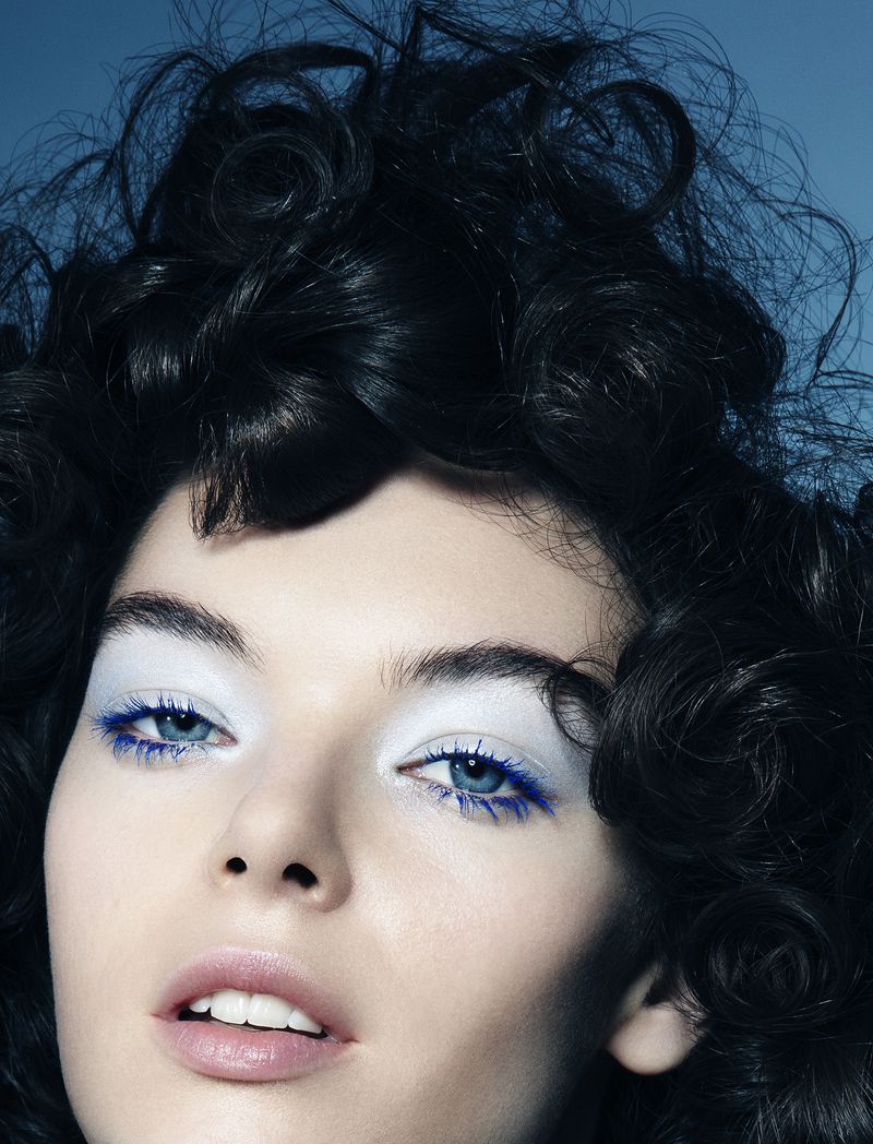 Headshot of a woman with dramatic makeup including white eyeshadow and blue eyeliner and mascara, along with dark curly hair.