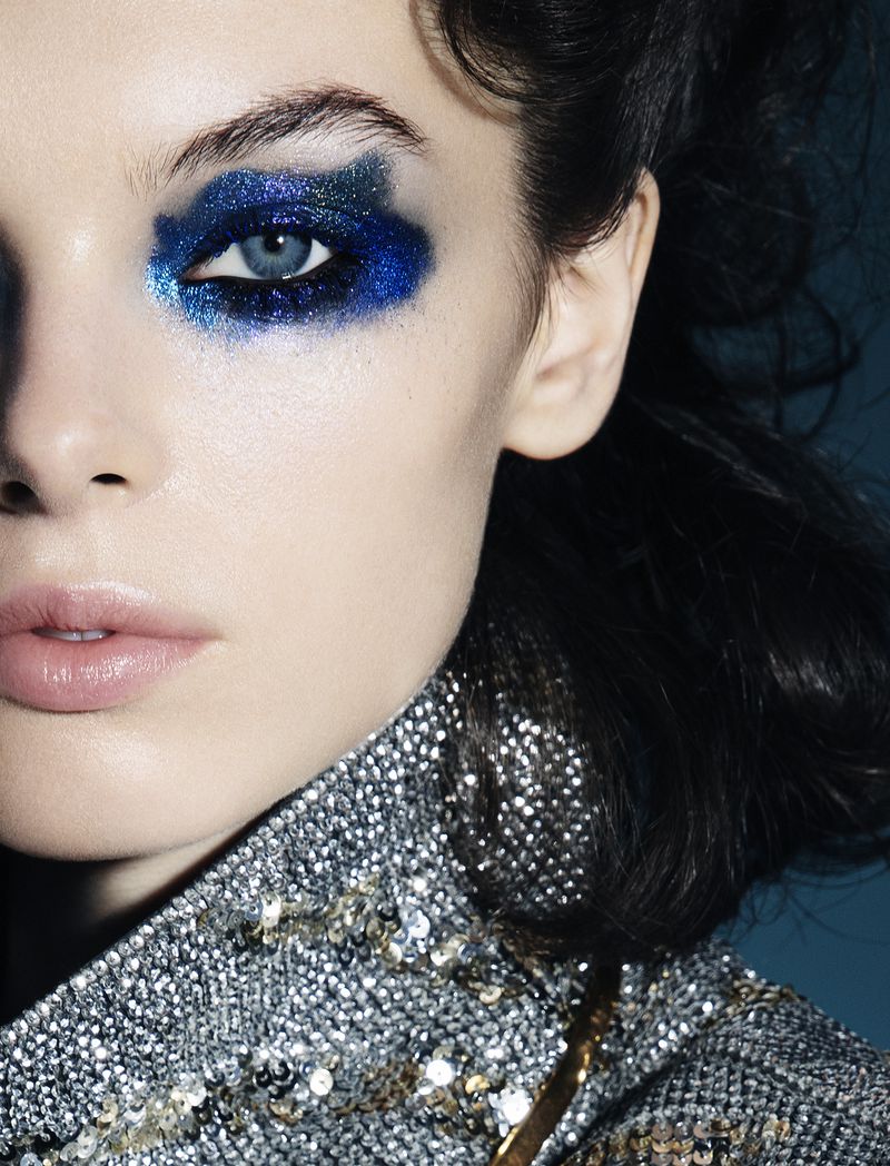 Close-up of a young woman's face with bold, sparkling blue eyeshadow, glossy lips, and silver sequined clothing against a dark blue background.