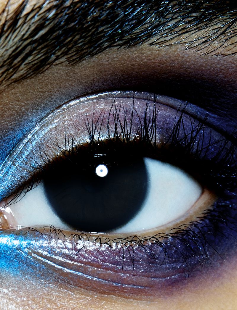 Close-up photograph of an eye with blue and purple eyeshadow, focusing on the details of the eyelashes and skin texture.