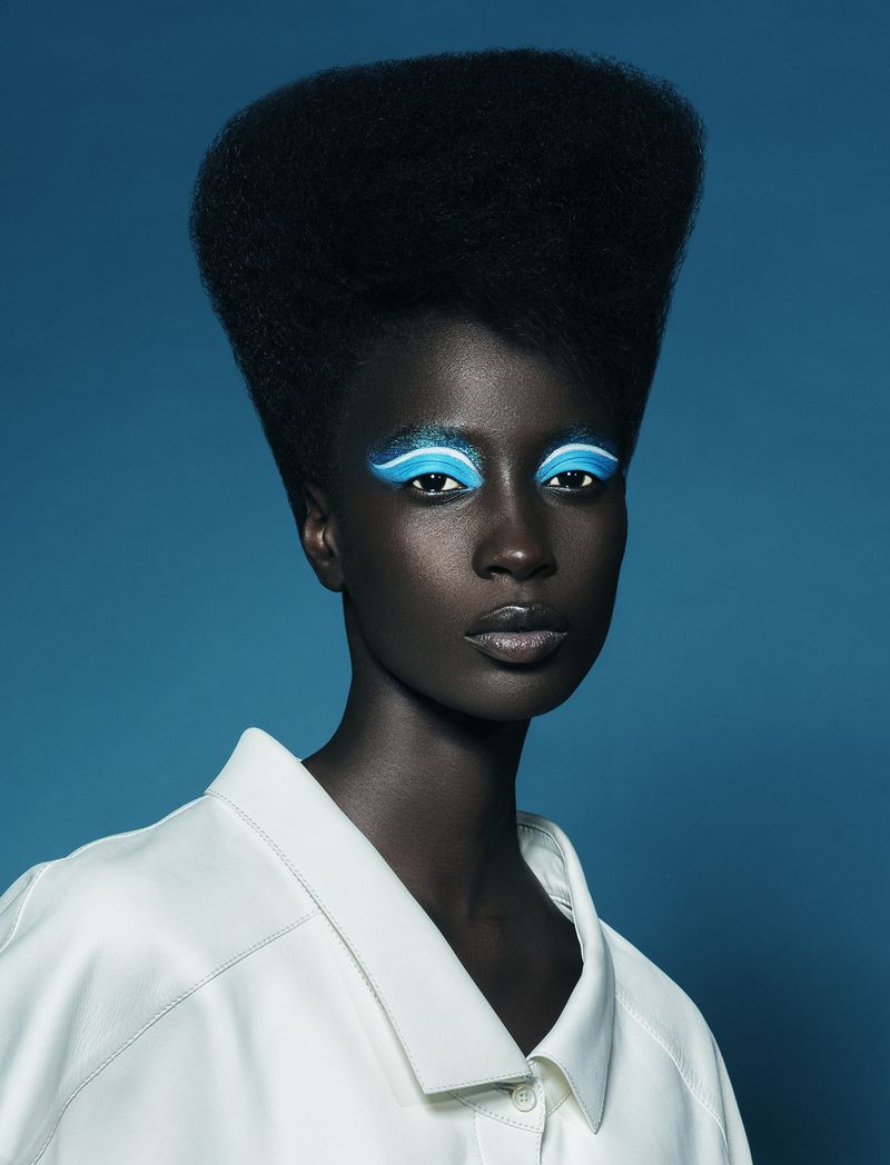 Close-up portrait of a dark-skinned woman with a tall, afro hairstyle, wearing bright blue eyeshadow and a white shirt against a blue background.