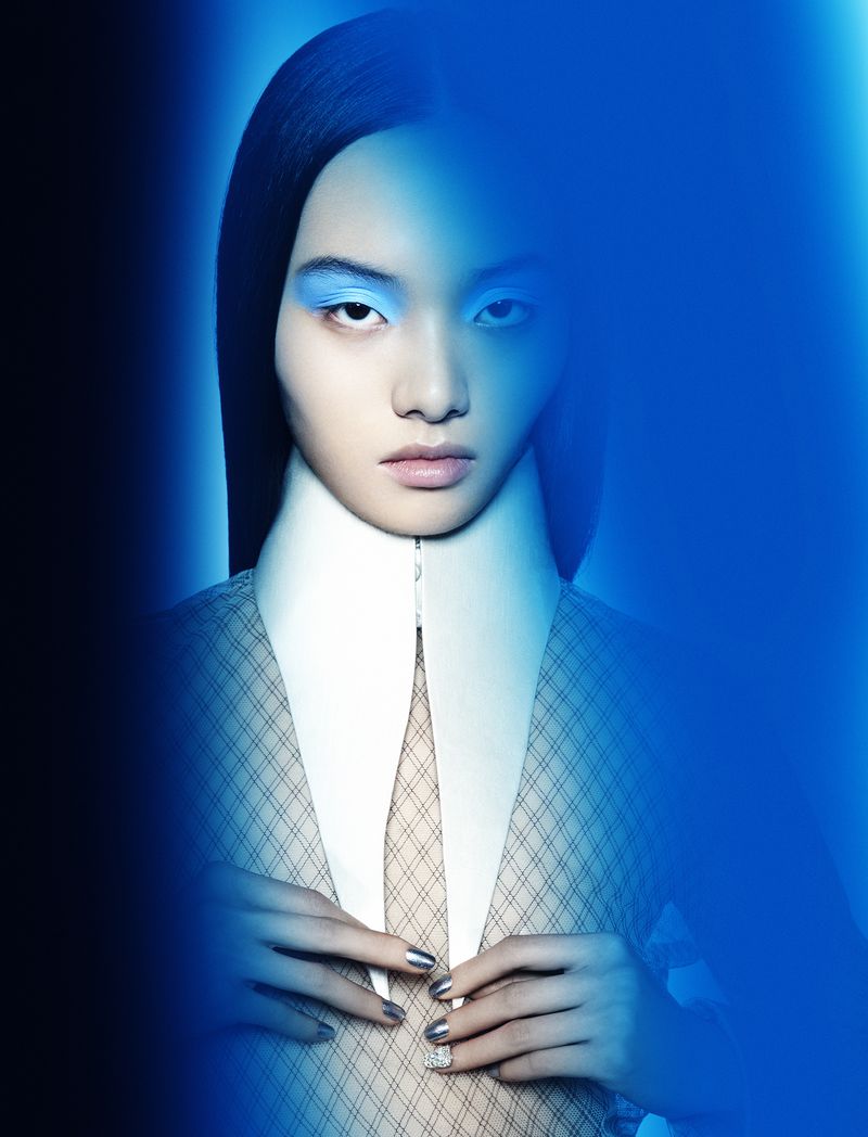 Eye-level studio portrait of a young woman with bright blue eyeshadow, a white collar, hands near buttons of a patterned blazer, and blue light.