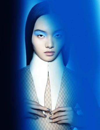 Eye-level studio portrait of a young woman with bright blue eyeshadow, a white collar, hands near buttons of a patterned blazer, and blue light.