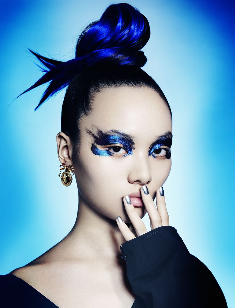Eye-level portrait of an adult female with blue hair and makeup, wearing gold earrings, set against a soft blue background.