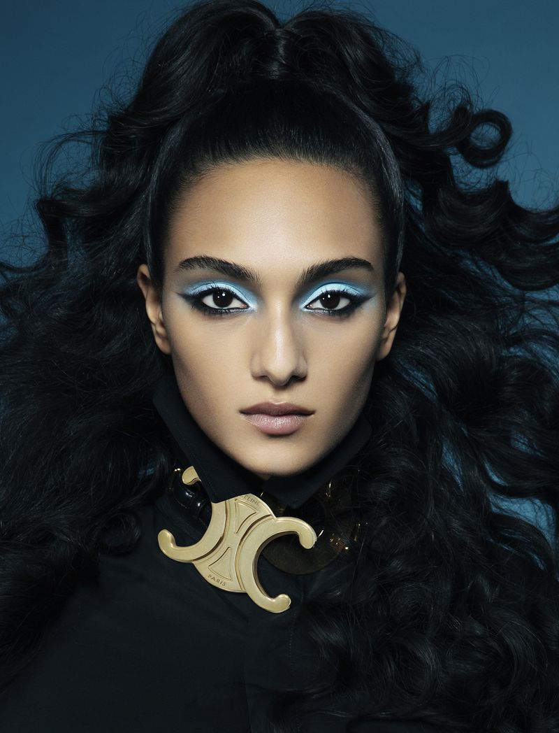 Close-up of a brunette adult female wearing statement necklace and light blue eye makeup against a dark teal background in studio.