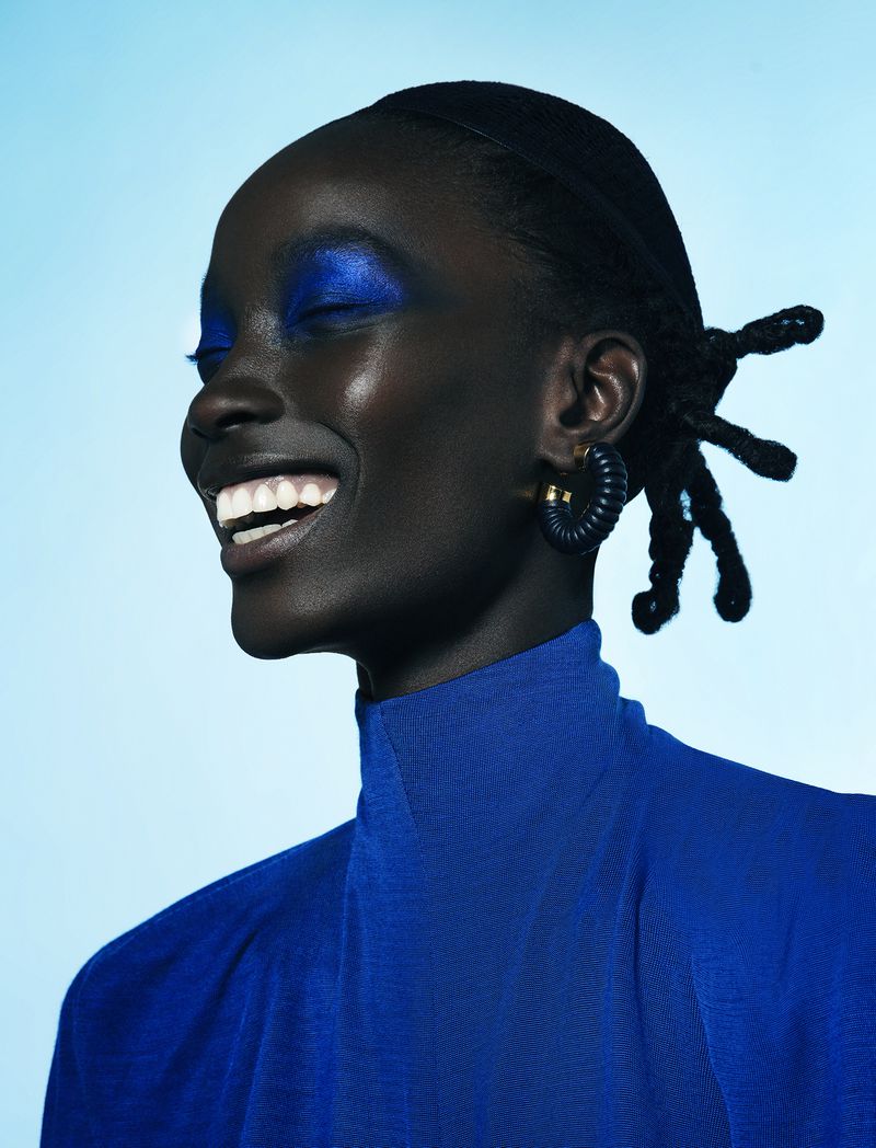 Close-up portrait of a smiling Black woman with closed eyes, wearing bright blue eyeshadow and a matching top against a light blue backdrop.