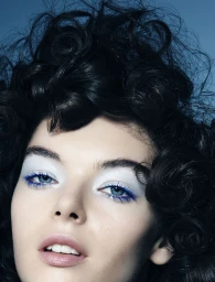 Close-up portrait of a woman with voluminous dark curly hair, blue mascara, and pale blue eyeshadow against a blue backdrop.
