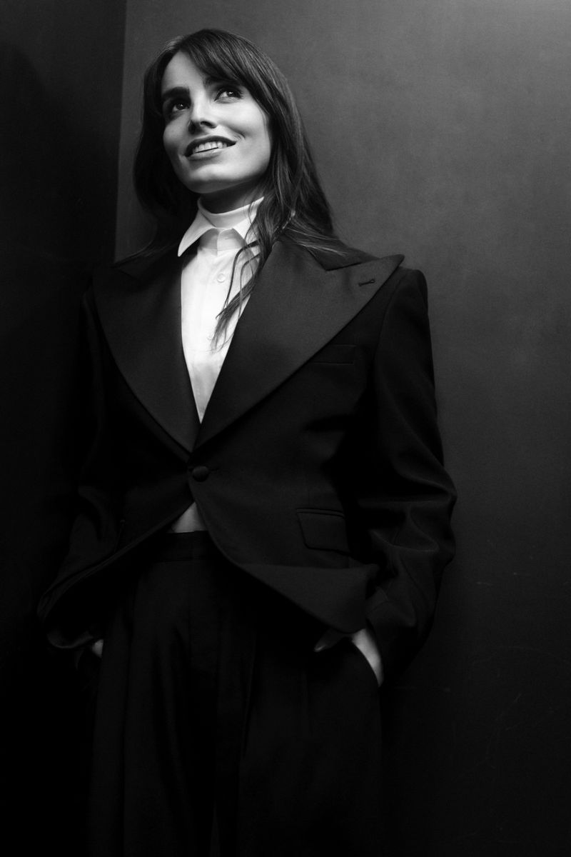 Black and white portrait of a woman in a black tuxedo, looking up and to the right. The background is a solid dark gray.
