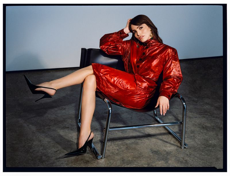 Adult woman with dark hair sits in a black chair wearing a red jacket and skirt with black heels in a studio setting.