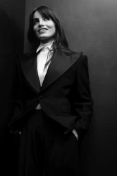 Portrait of Lux Pascal wearing a dark tailored suit jacket and white shirt against a dark studio background.