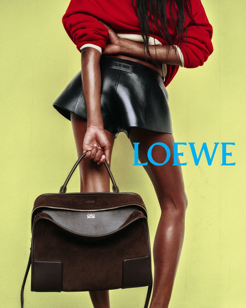 Fashion shot of a woman in a red sweater and black leather shorts holding a brown suede handbag against a yellow backdrop.