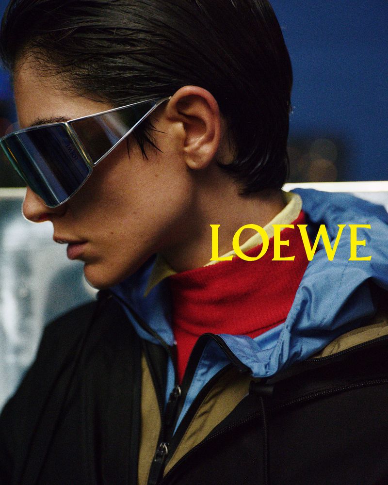 Fashion shot of an adult with futuristic sunglasses, red turtleneck, and layered jacket with the Loewe logo in front of a blue background.