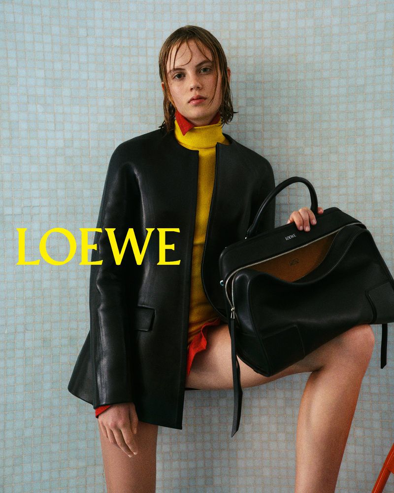 Editorial fashion shot of a young woman in a black jacket, yellow turtleneck, and holding a black leather bag against a tiled wall.