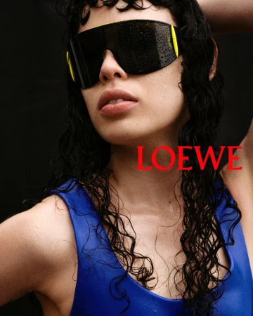 Model wearing dark visor sunglasses and a blue top against a dark background with red Loewe typography