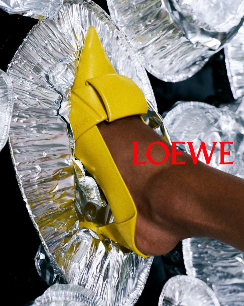 Close-up of a yellow high-heeled shoe on a dark-skinned model, posed against a background of crumpled silver foil.