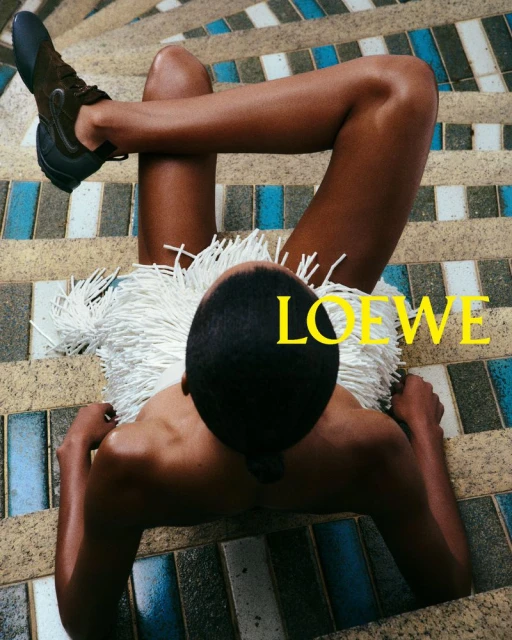 High-angle view of an adult with dark skin and short black hair lying on a tiled staircase wearing brown shoes and a fringed dress.