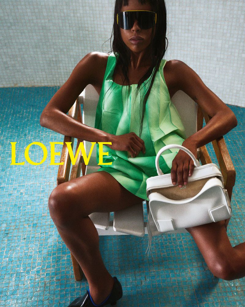 Fashion shot of a woman in a green dress, sunglasses, and black shoes sitting in a chair, holding a Loewe white handbag.