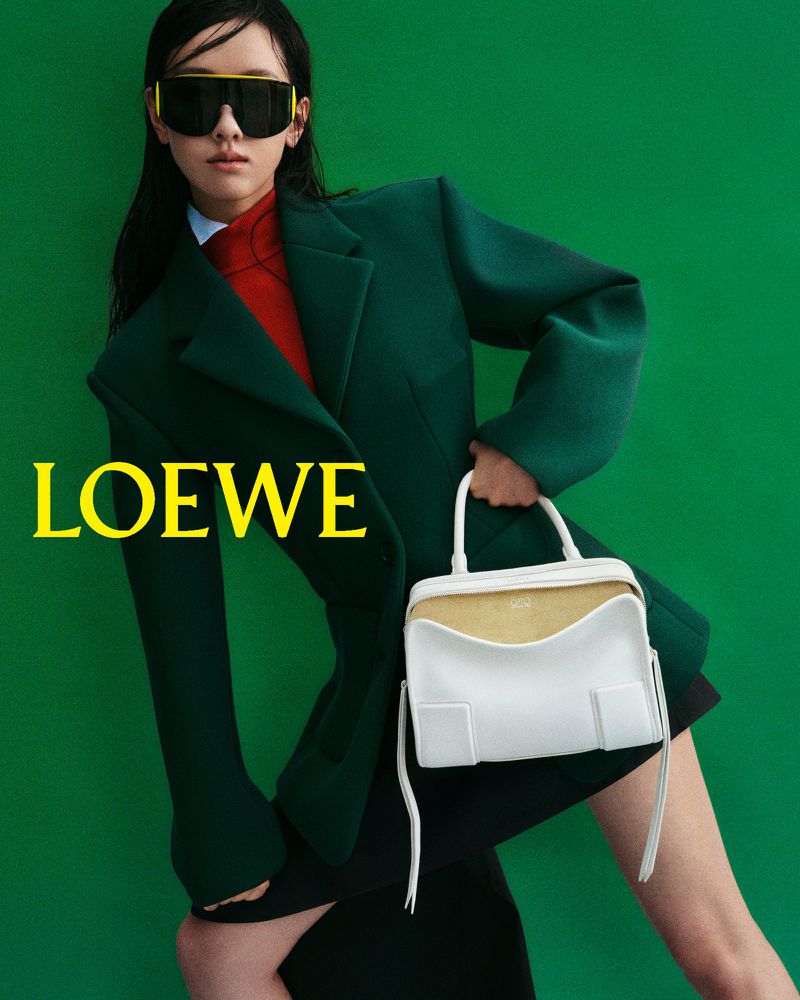 Fashion shot of a model wearing a green blazer and sunglasses, holding a white handbag, posing against a green studio backdrop.