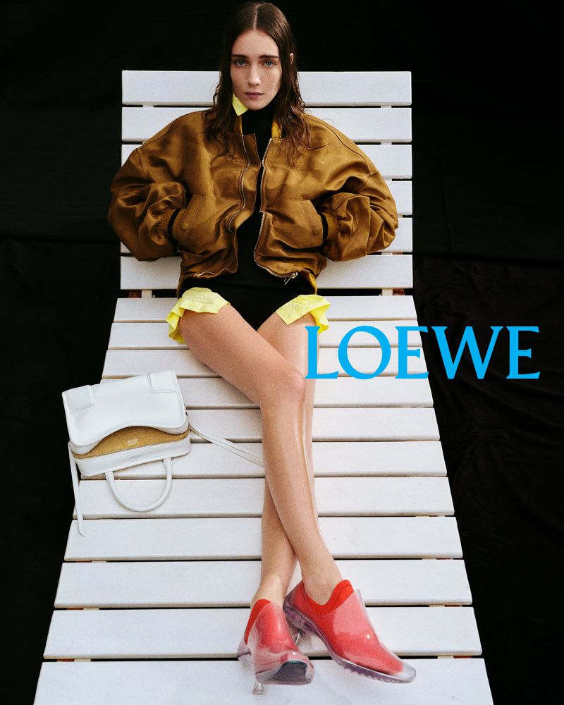 Fashion editorial shot of a young adult female model wearing a bomber jacket and red shoes, sitting on a white slatted surface.
