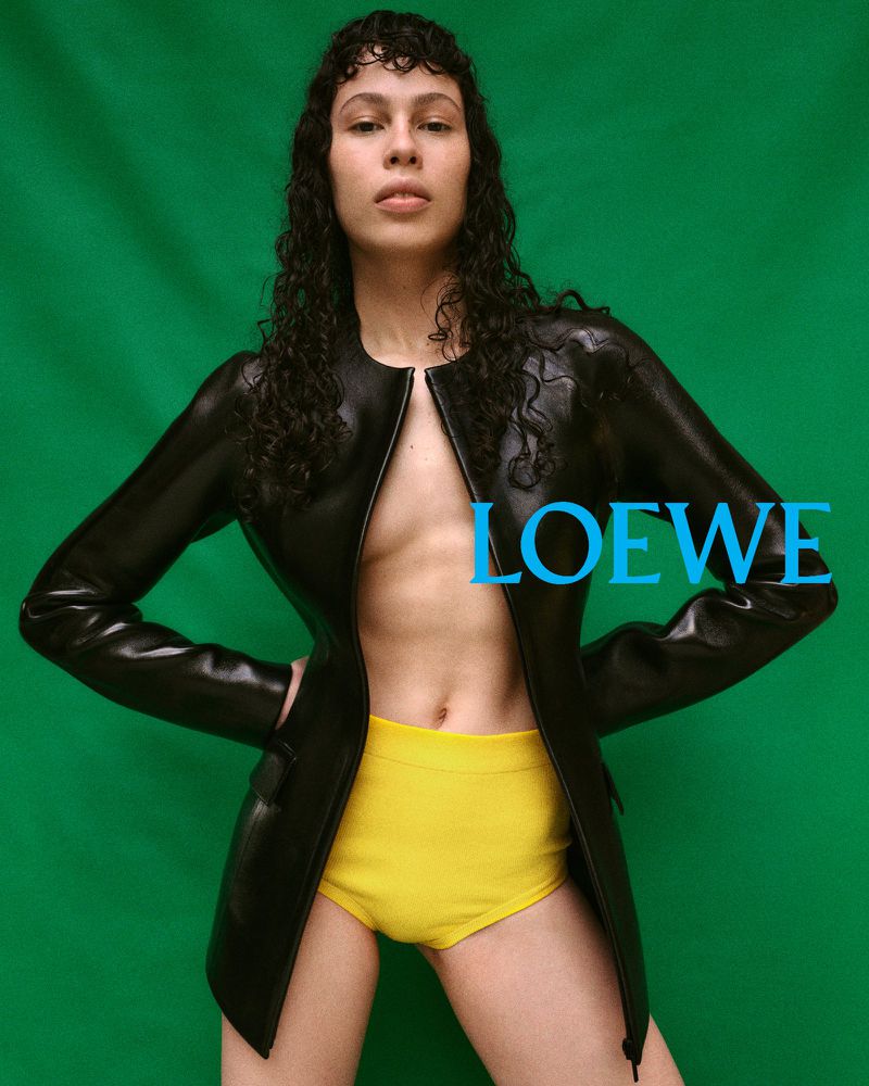 Woman poses in black leather jacket and yellow briefs against a green backdrop, showcasing fashion and style in a studio setting.