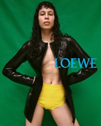Model with dark curly hair wearing a black jacket and yellow shorts against a green background