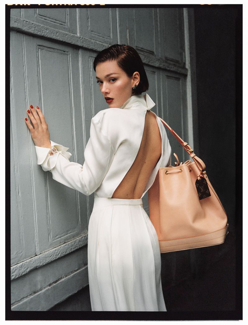 Indoor fashion shot of a woman in a white dress with an open back, carrying a large tan purse and standing next to a blue door.