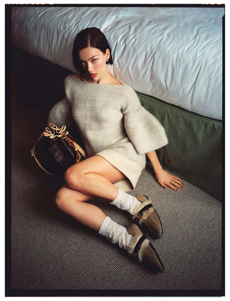 A woman in a light-colored knit sweater and skirt sits on a grey carpet next to a bed with white linens and a decorative bag.