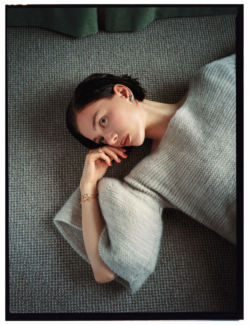 Woman with dark hair lies on a gray textured surface wearing a gray sweater and gold bracelet, looking at the camera with a serious expression.