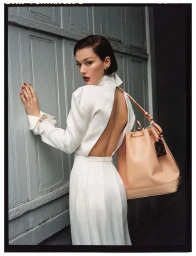 Woman in white backless dress wearing a large peach leather bucket bag against a grey panelled door