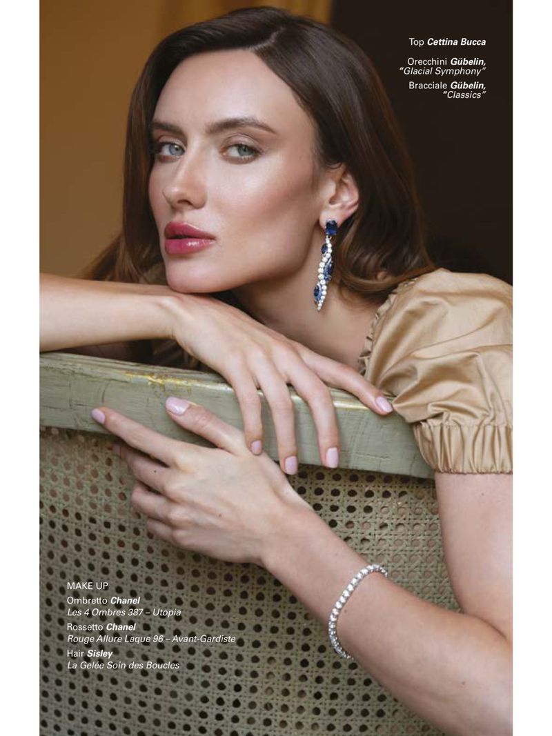 Close-up portrait of a woman with long brown hair wearing chandelier earrings and a tennis bracelet, resting her hands on a carved chair.