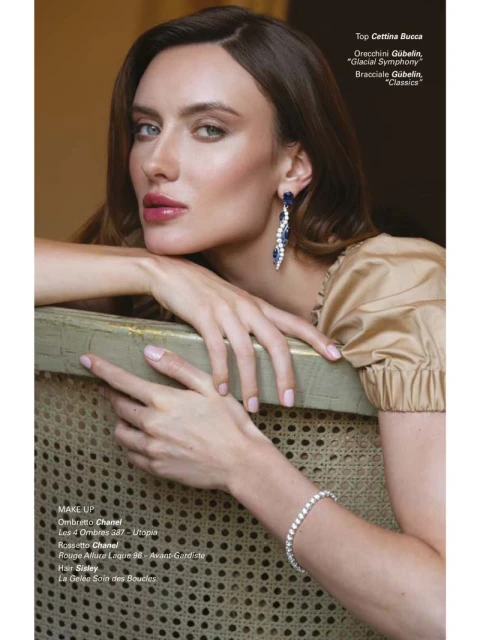 Close-up portrait of a woman with long brown hair wearing chandelier earrings and a tennis bracelet, resting her hands on a carved chair.