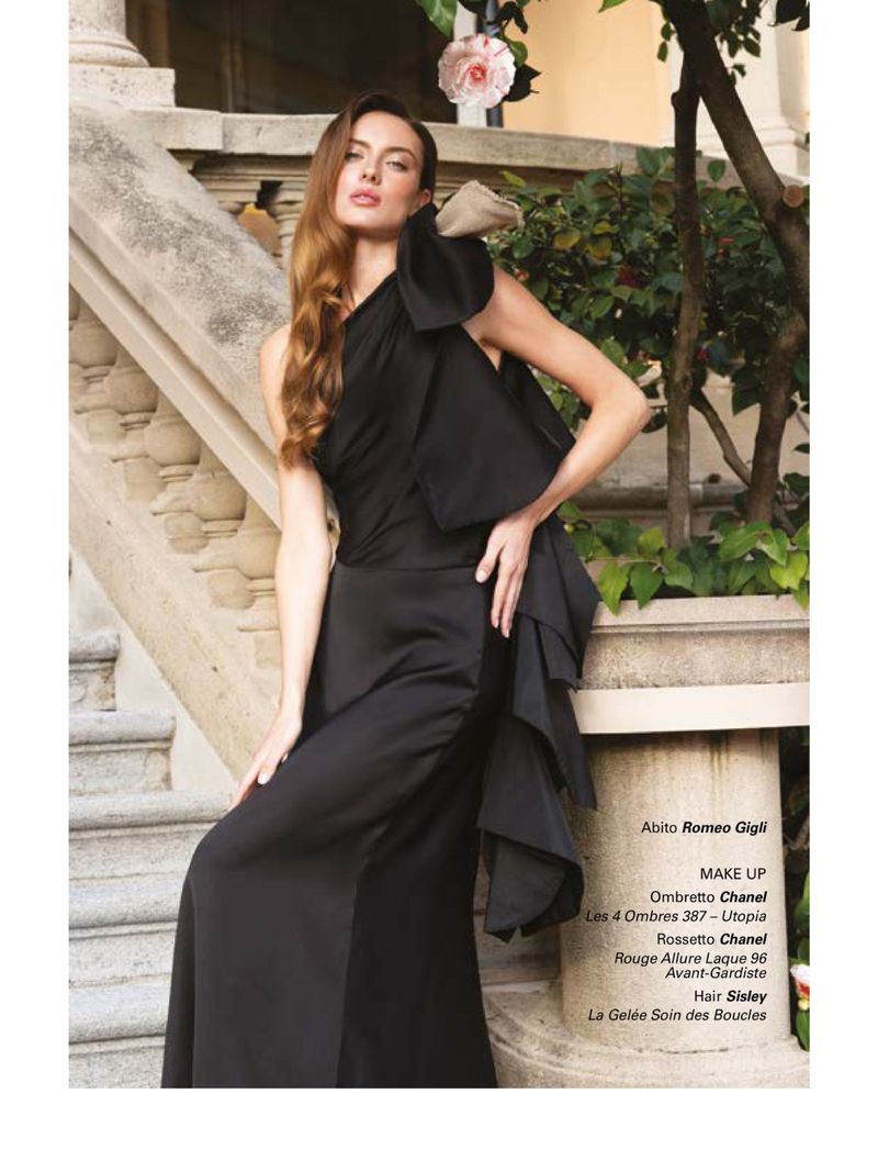 Elegant woman in black one-shoulder gown posing on stairs. Fashion editorial shot with architectural and floral background.