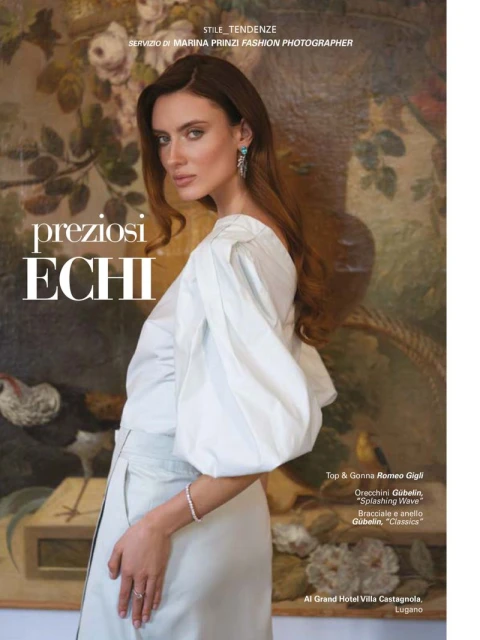 Fashion editorial shot: A brunette woman poses indoors wearing an elegant white blouse and skirt, showcasing designer jewelry in front of a classic painting.