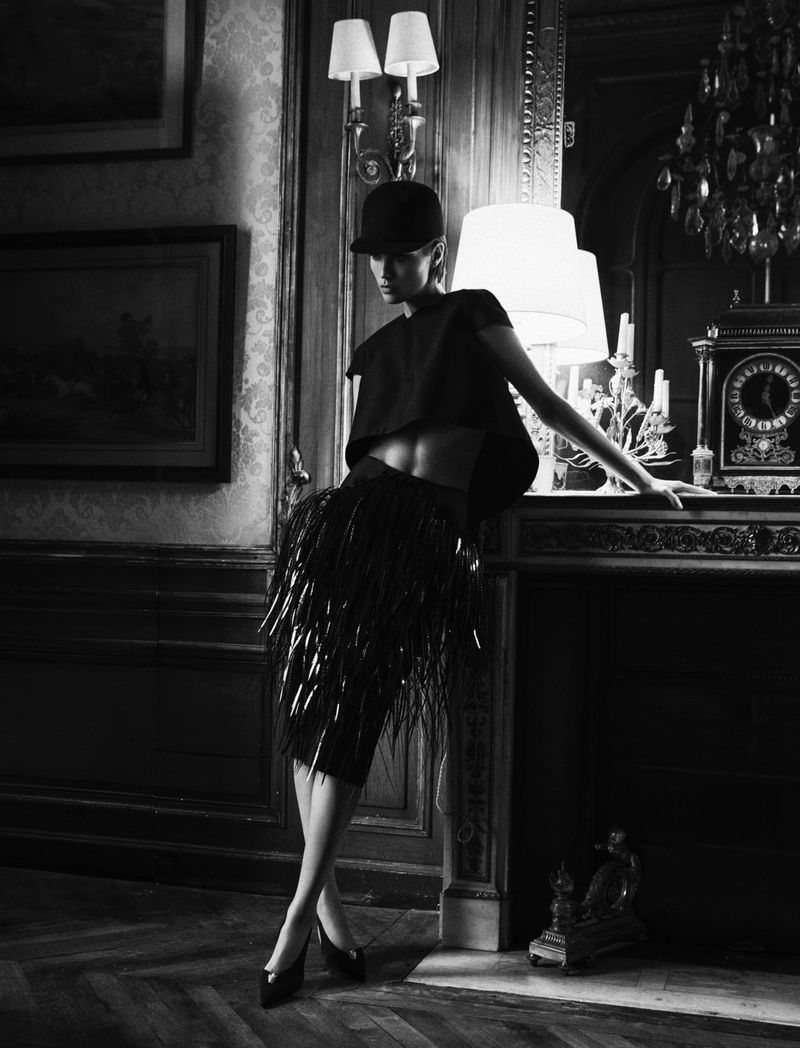 Fashionable woman in cap and fringed skirt poses near fireplace with decorative elements in an elegant, moody, black and white interior.