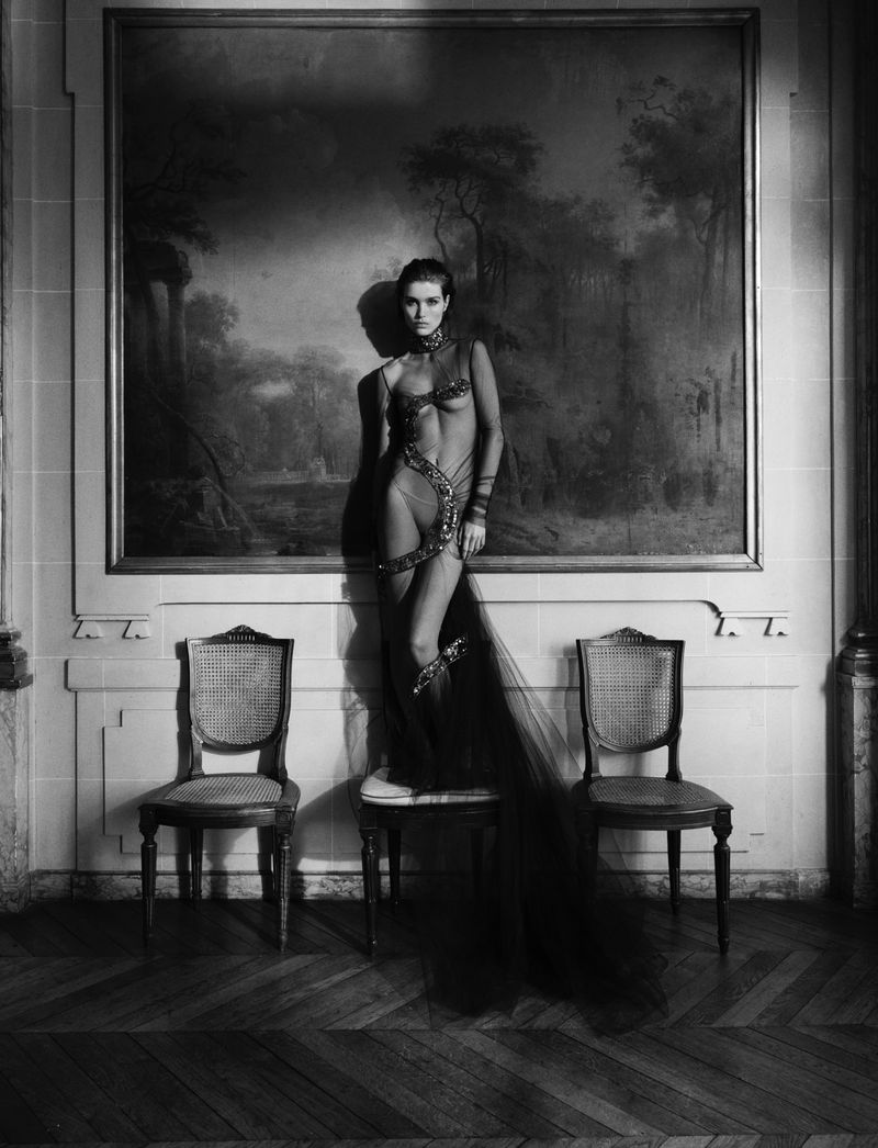 Monochrome full shot of a woman in ornamental attire leaning on a small table, flanked by chairs, with a large painting behind her.