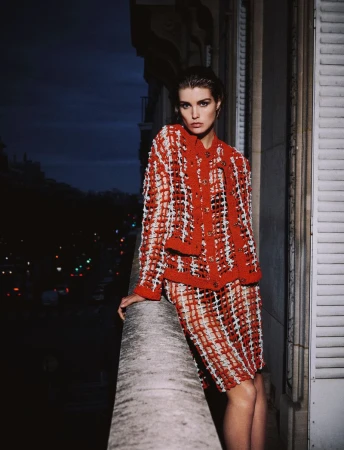 Fashionable adult woman poses on a balcony at night wearing a red and white tweed skirt suit, with a cityscape visible in the distance.