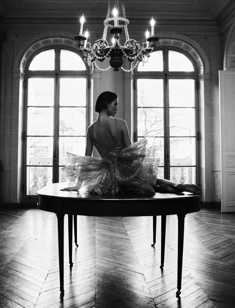Black and white photo of an adult woman in a tutu seated on a table, looking away in a room with high ceilings and large windows.