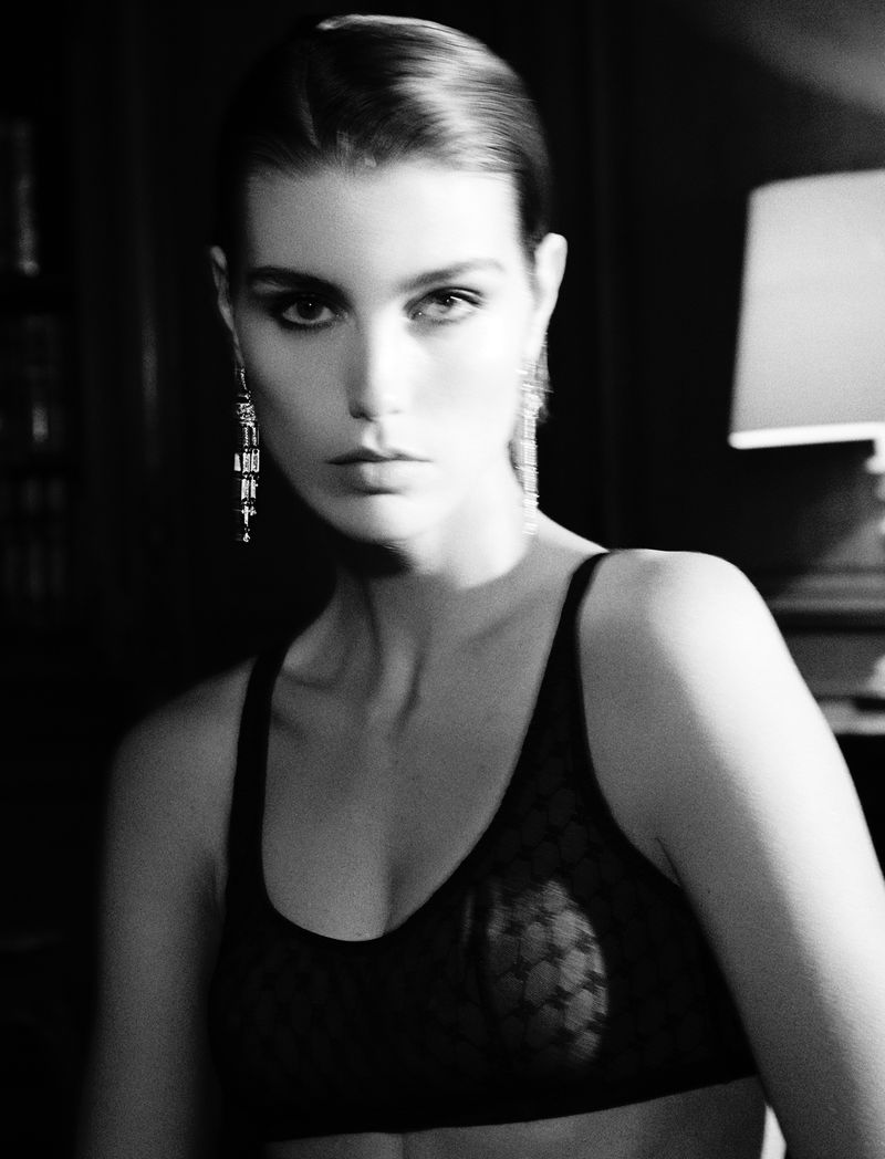 Black and white portrait of a woman with dark hair and dangling earrings, wearing a lace bra in an indoor setting with soft lighting.