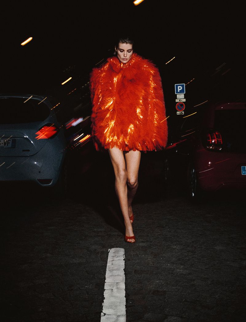 An adult female with fair skin, wearing a red, shiny coat and red high heels, walks in a street at night with cars and signs in view.