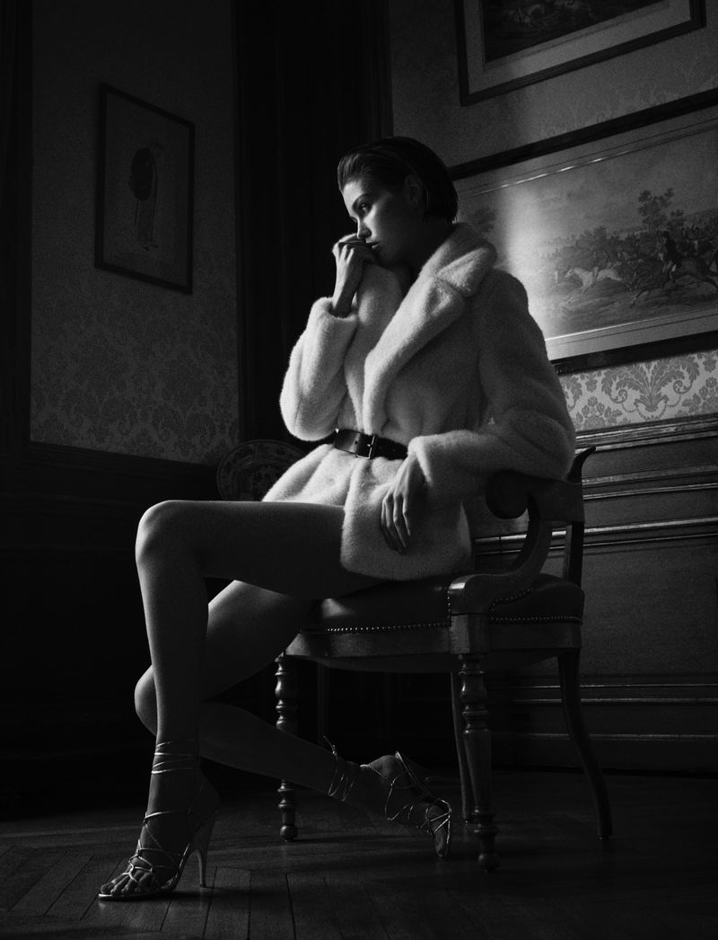 Model Luna Bijl seated on a classic chair wearing a white coat and heels in a dark room