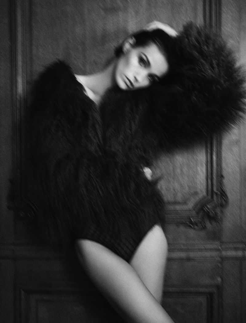 Elegant woman in a dark furry coat poses against a paneled door, exhibiting high fashion style with a touch of vintage flair in a studio.